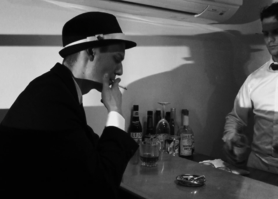 A screenshot from the film, showing a man in a fedora holding a drink, sitting at a bar