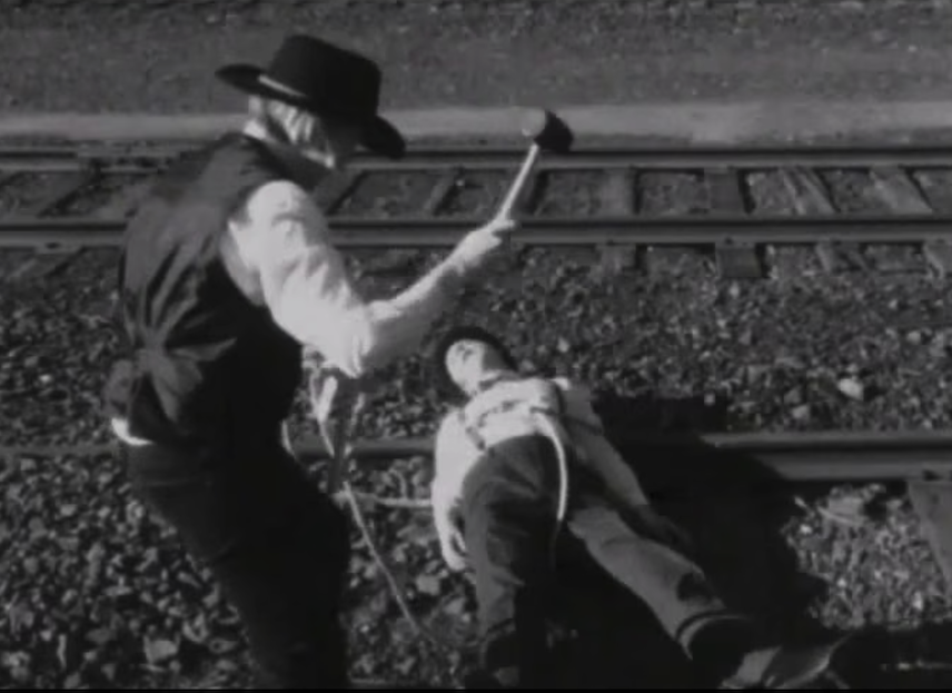 A screenshot from the film, showing a man tied to railroad tracks, and a villain threatening him with a hammer